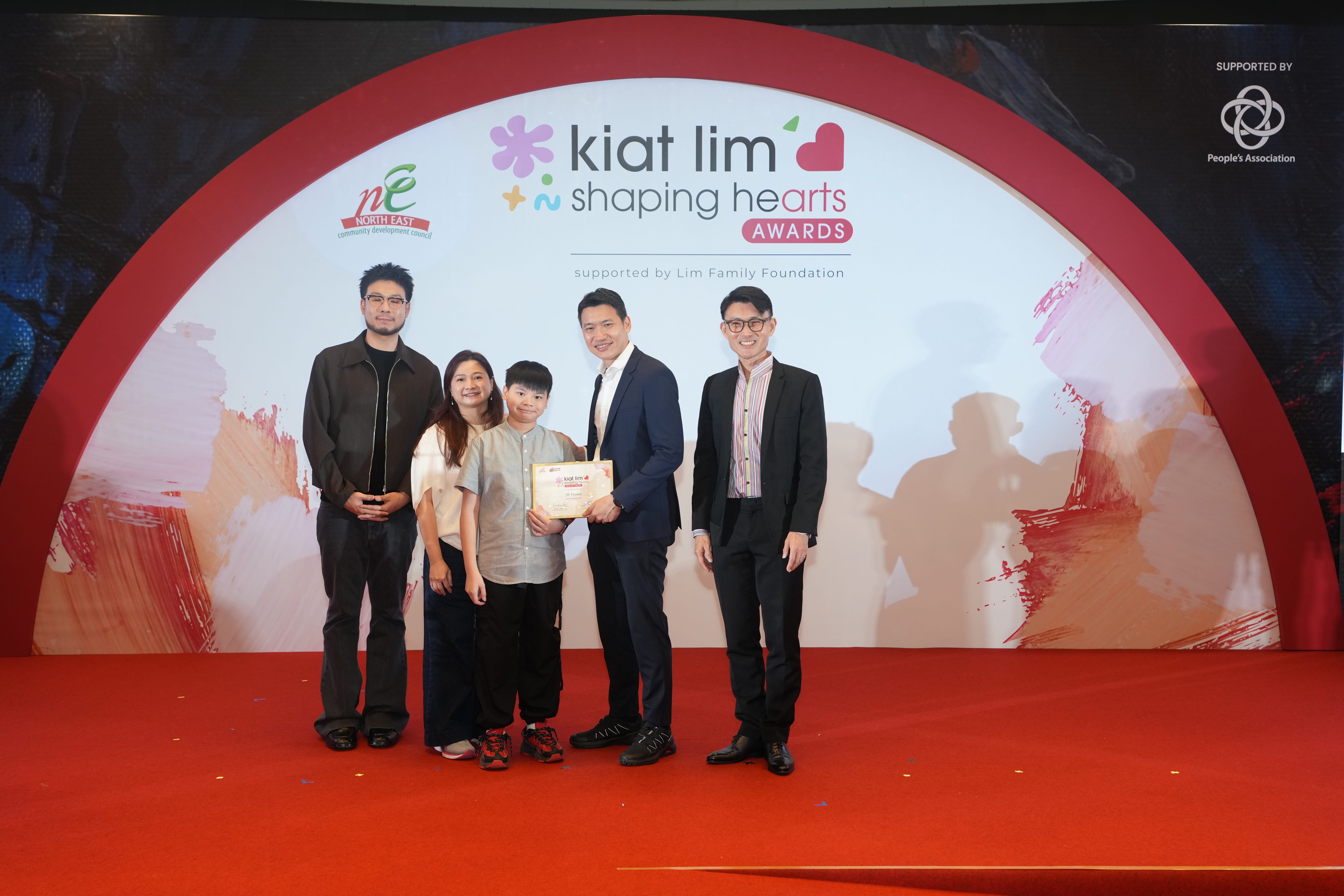 Award recipient standing on stage with the presenter, holding their certificate and posing for a group photo during the award ceremony, with a backdrop reading ‘Kiat Lim-Shaping Hearts Awards'.